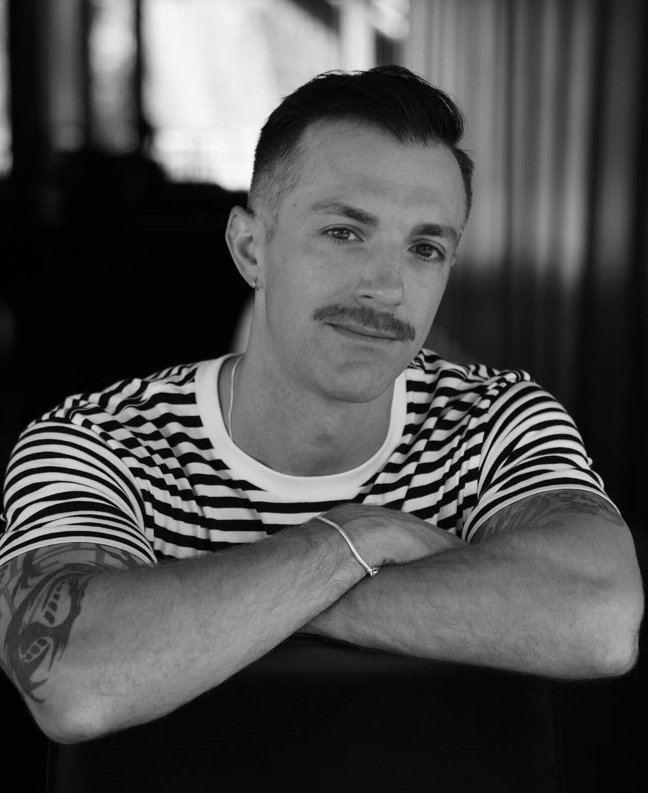 Sean Horlor, documentary director and filmmaker, in a portrait from Sitges Film Festival in Spain, featuring a smiling man in a striped shirt with a moustache and tattoos visible on his arms.